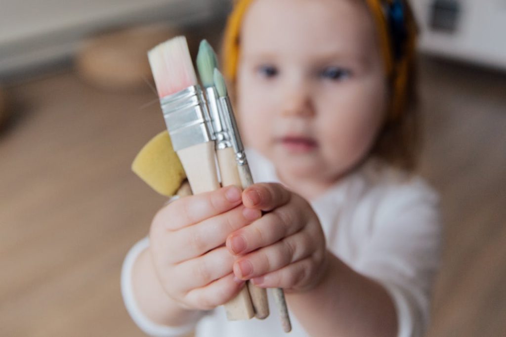 A cute toddler enjoying creativity by holding several art brushes indoors.